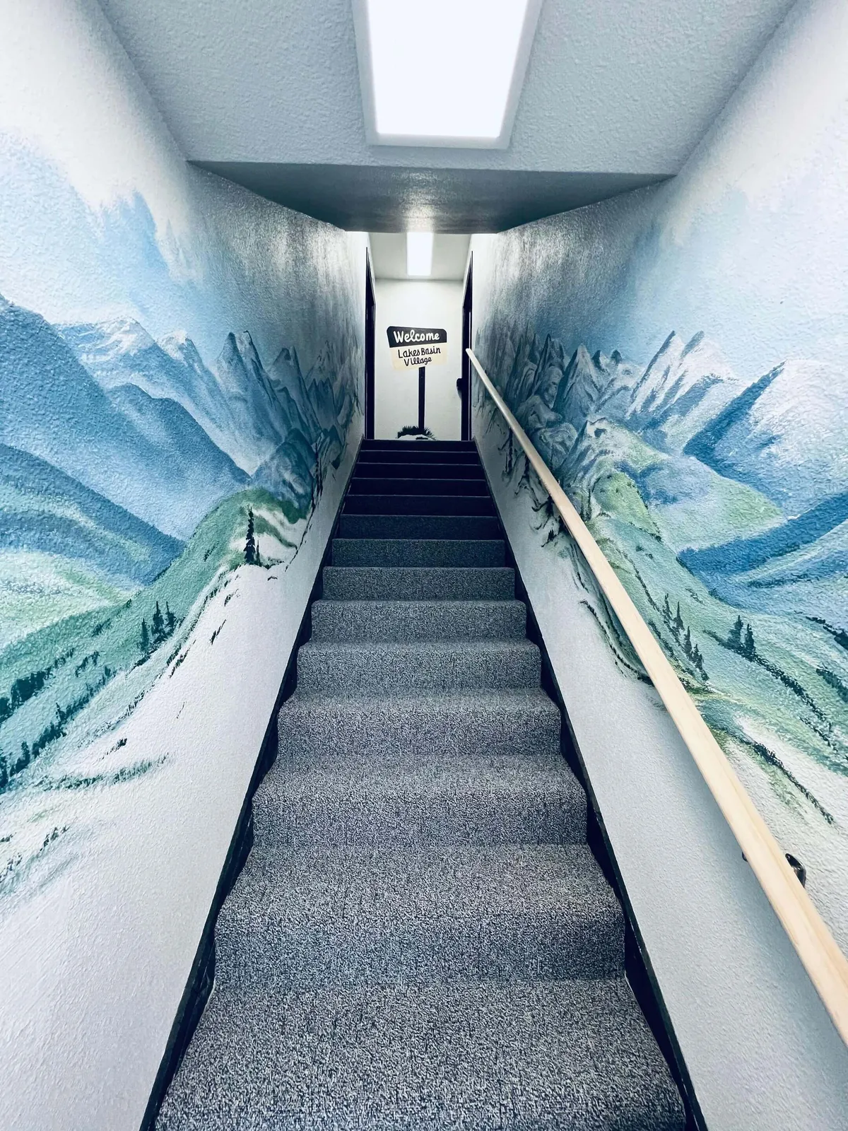 A warmly lit entryway featuring a prominent mural on the wall, a staircase, and a visible 'welcome' sign. The space feels inviting and artistic, setting the tone for a welcoming arrival into the condo and highlighting the sense of community and individuality within the property.