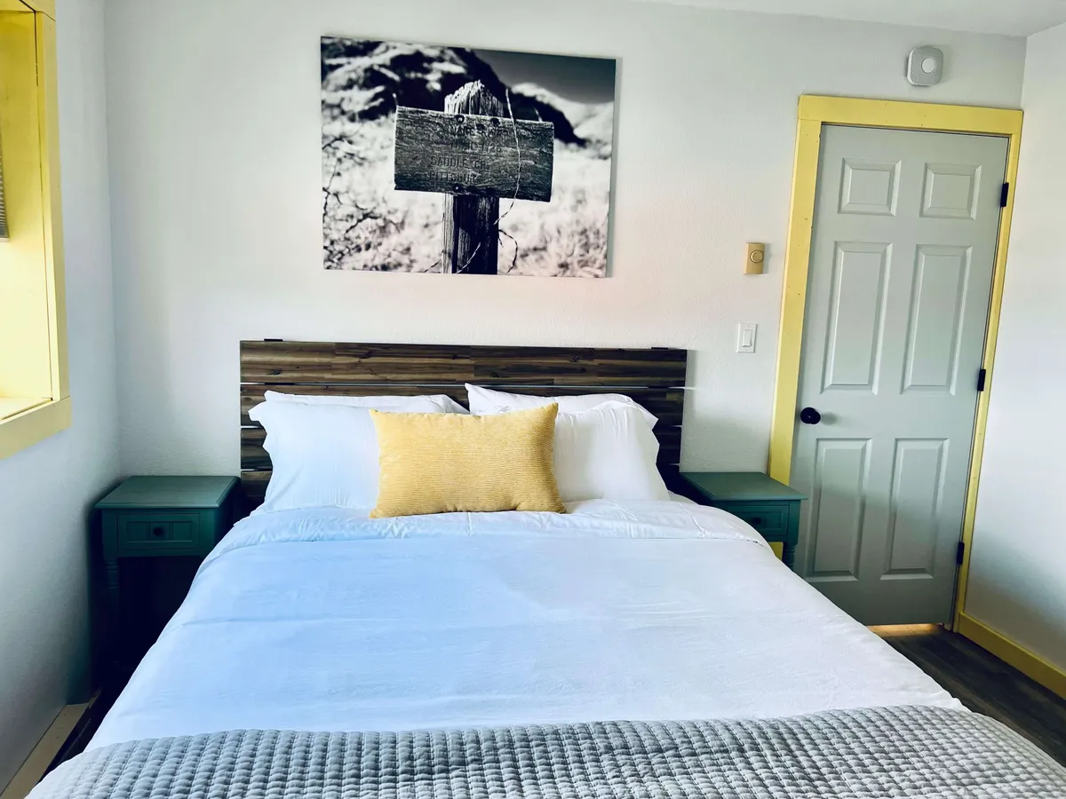 A cozy bedroom scene with a comfortable queen bed, white linens, a yellow accent pillow, and a wooden headboard. The green nightstand and a framed photograph on the wall add warmth and a touch of personality. The space is bathed in gentle natural light, enhancing its tranquil feel.