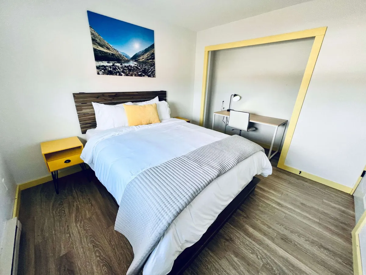 A bright and inviting bedroom featuring a plush queen bed dressed in white linens and accented with a cheerful yellow pillow. A yellow nightstand sits beside the bed, while a landscape artwork hangs above, and natural sunlight fills the room, creating a serene atmosphere.