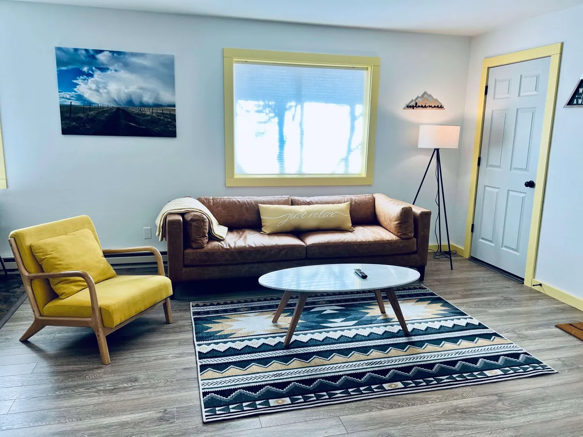 The image showcases a cozy living room with a plush couch, a cheerful yellow chair, a stylish coffee table, and a colorful rug. The space is accentuated by wall art and bathed in natural light, creating an inviting atmosphere ideal for relaxing, reading, or socializing.