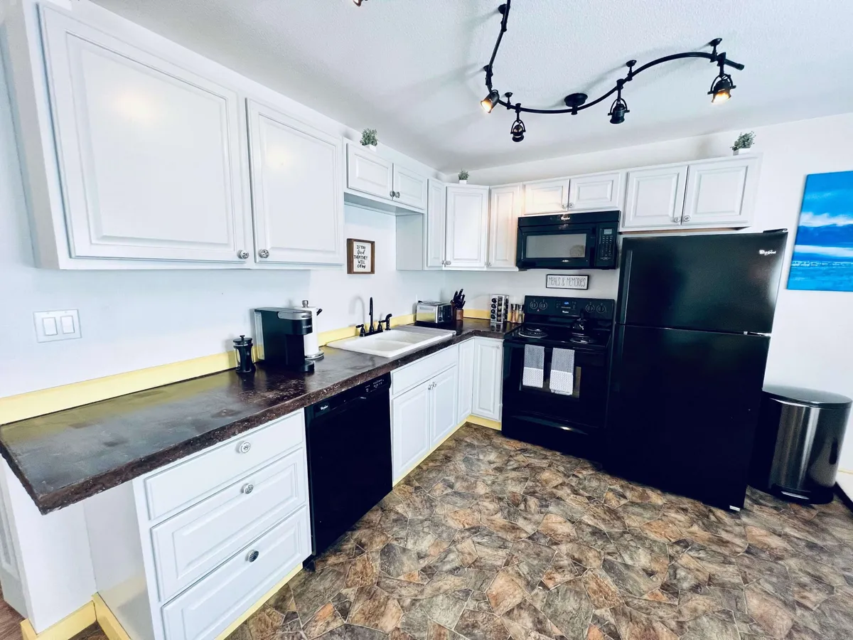 This image presents a bright, functional kitchen space featuring crisp white cabinetry, sleek black appliances, and a spacious countertop with a sink. The scene conveys a welcoming environment for cooking, socializing, and sharing meals, with enough room for family or friends to gather.