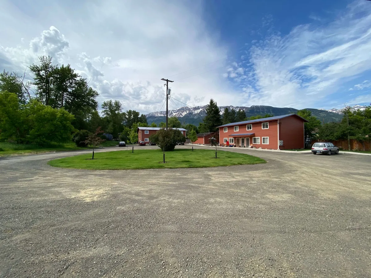 A picturesque exterior shot of red village-style buildings set against a lush green lawn and majestic mountain backdrop. The setting feels peaceful and communal, inviting outdoor relaxation and social interaction, with a clear sense of proximity to natural beauty.