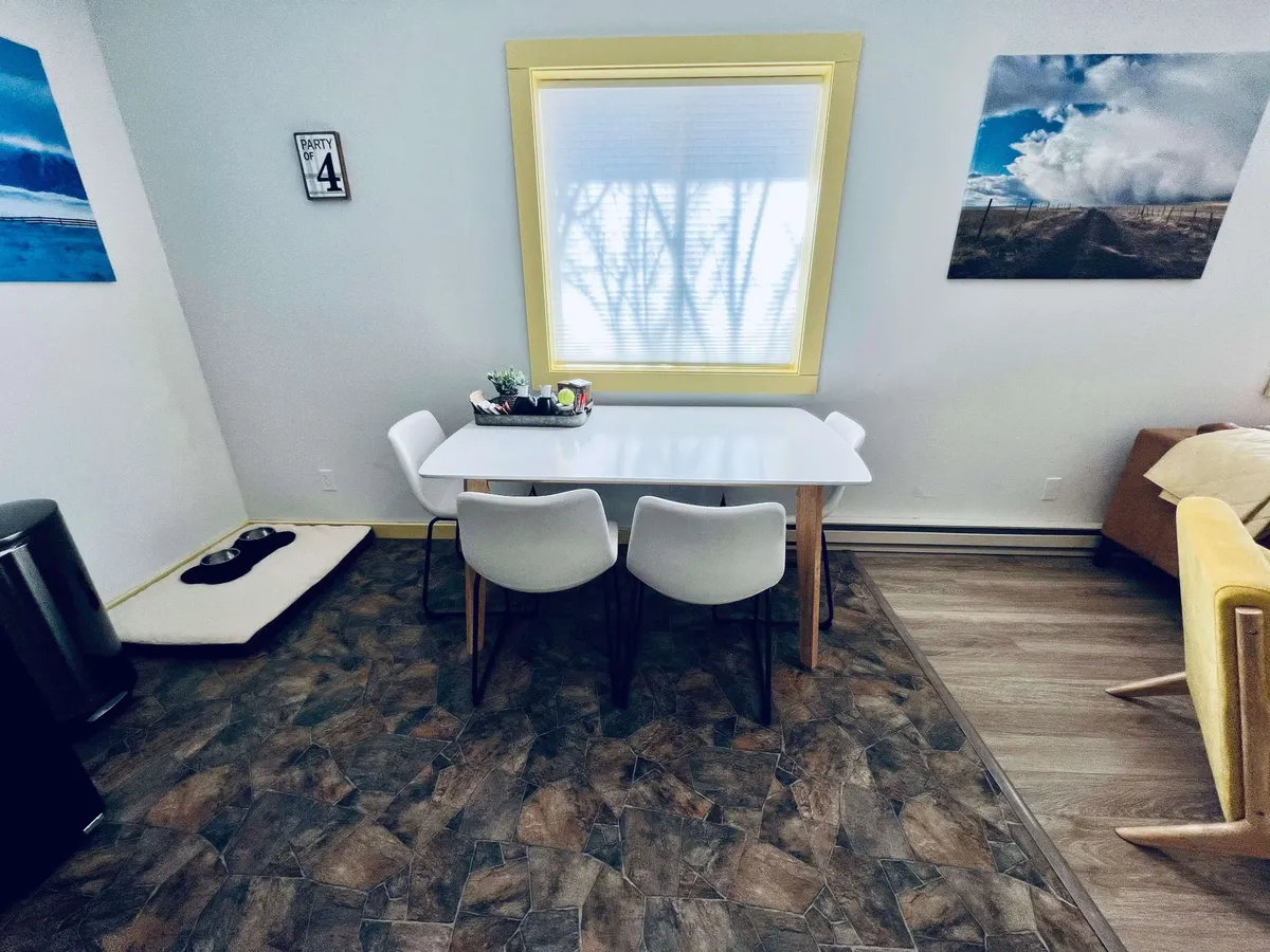 This photo highlights an open-concept interior where a dining table sits near a yellow-framed window. Soft light filters through, illuminating both the dining and living spaces. Decorative wall art and a comfortable sofa further connect the areas, reinforcing a sense of togetherness and warmth.