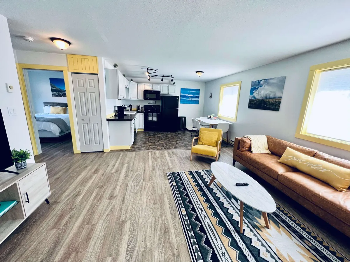 The image showcases a bright and inviting living space featuring a cozy sofa, a cheerful yellow chair, a dining table perfect for shared meals or game nights, a modern kitchen area, and tasteful artwork on the walls. Sunlight fills the open-concept room, creating a warm, welcoming atmosphere ideal for relaxing or entertaining.